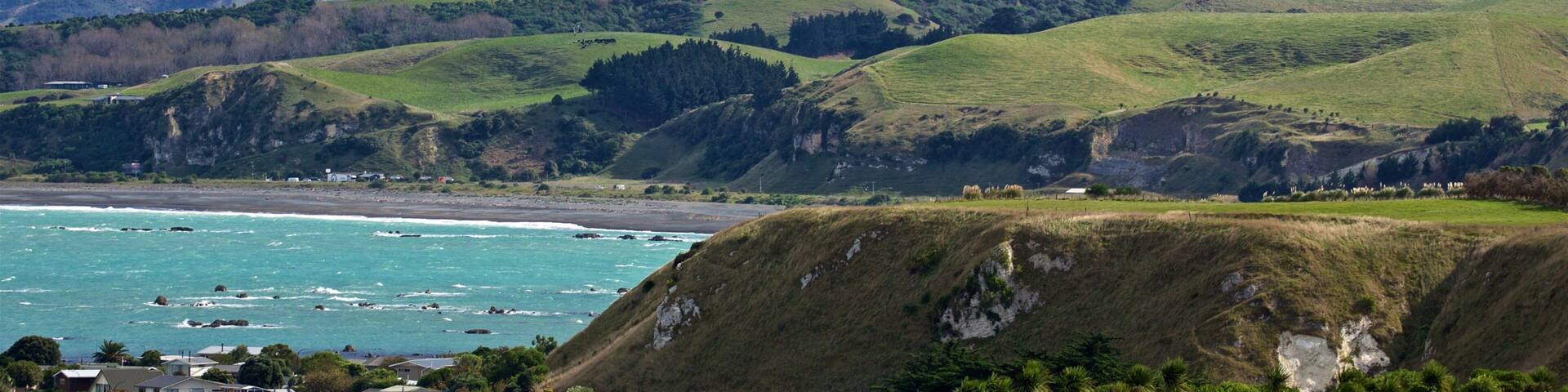 A coastal landscape with green hills, a beach, and snow-capped mountains in the background.