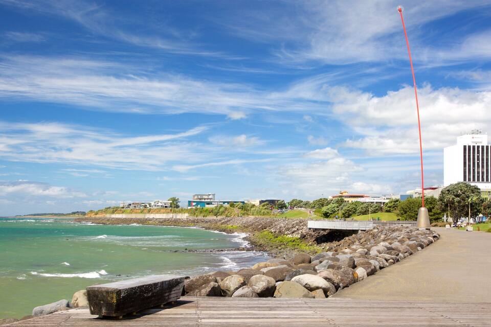 A coastal walkway with a rocky shoreline, ocean waves, and buildings in the background.