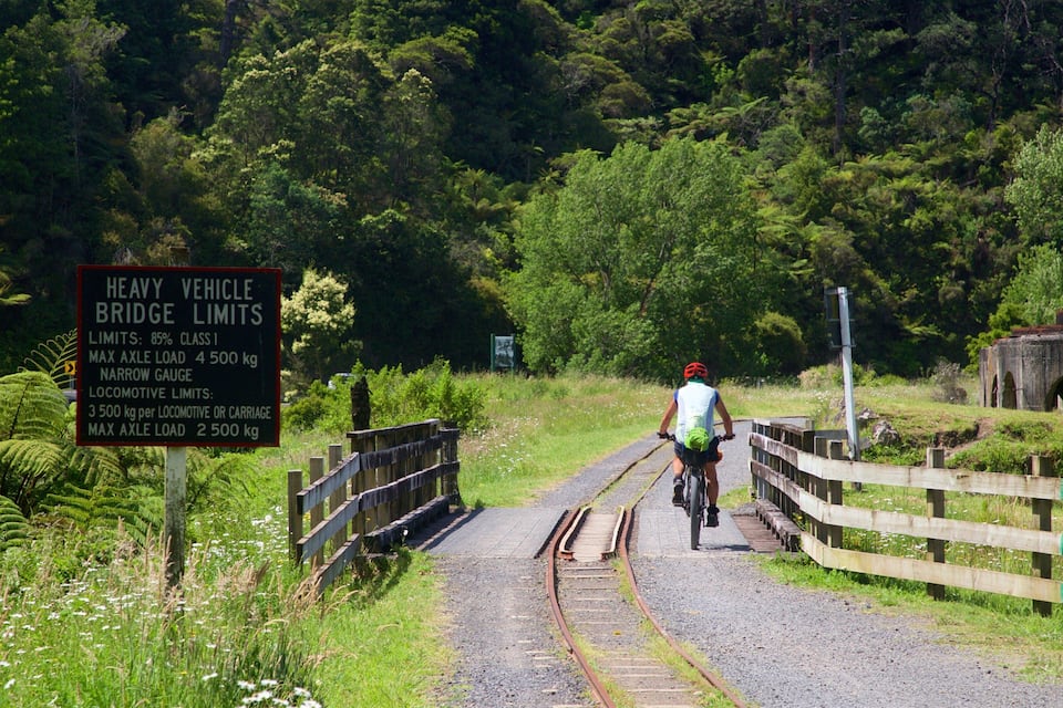 Un ciclista en un camino de grava junto a vías de tren, con un cartel que indica límites de peso para vehículos pesados.