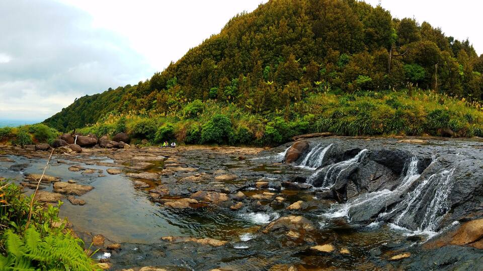A waterfall cascading over rocks in a lush, green valley with a forested hill in the background.