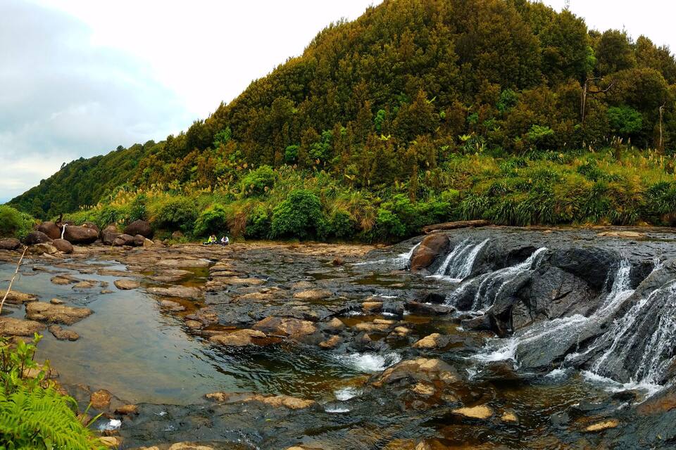 A waterfall cascading over rocks in a lush, green valley with a forested hill in the background.