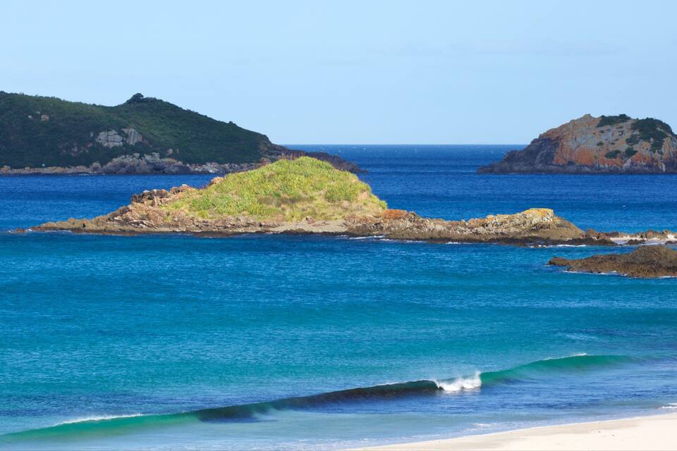 A beach with clear blue water, a sandy shore, and two rocky islands with vegetation.