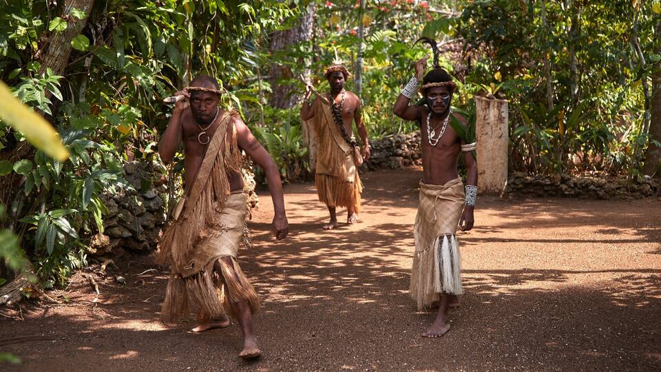 Three individuals in traditional attire walking on a dirt path surrounded by dense tropical foliage.