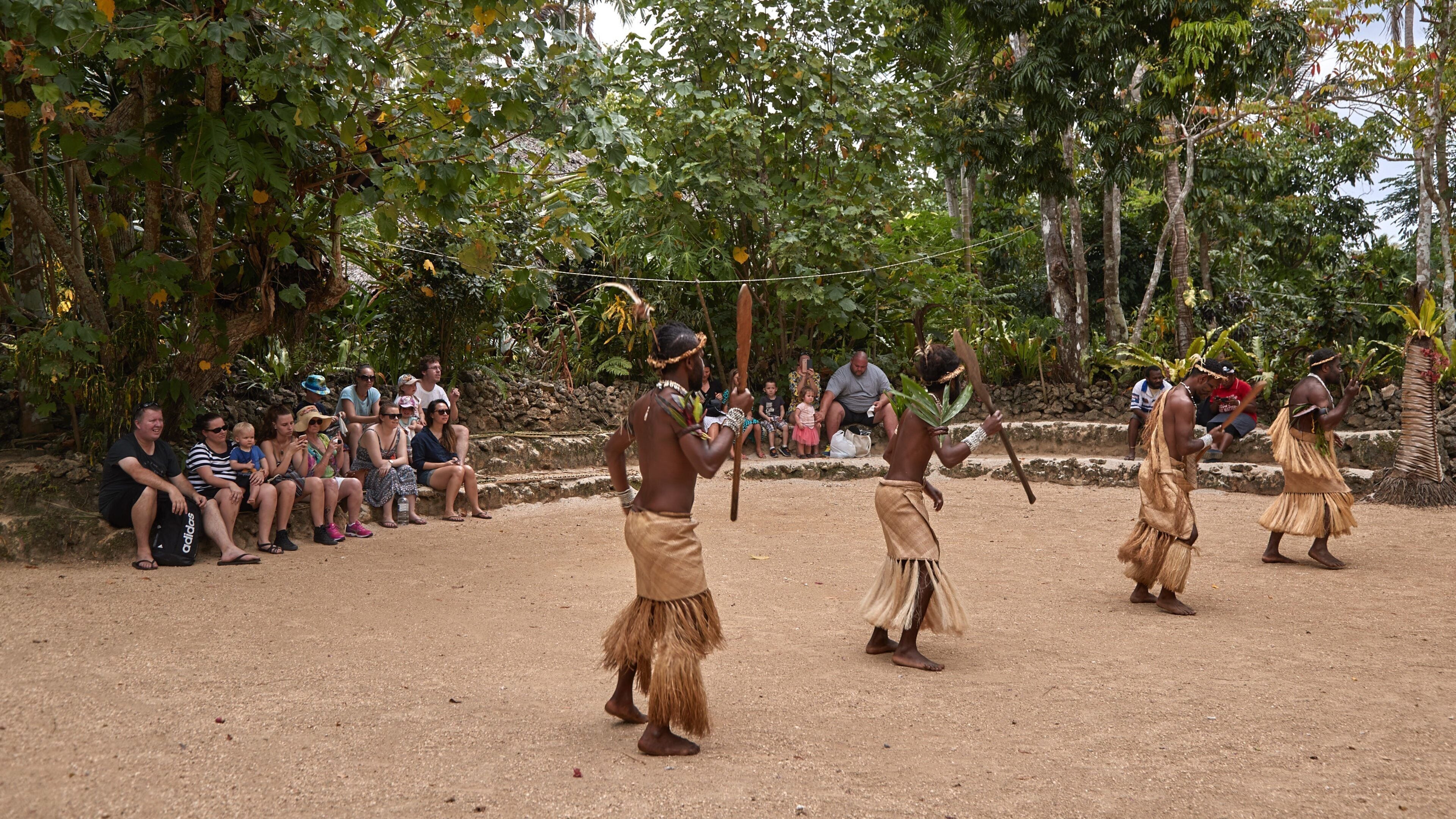A group of people in traditional attire performing a dance in an outdoor setting with a crowd watching.