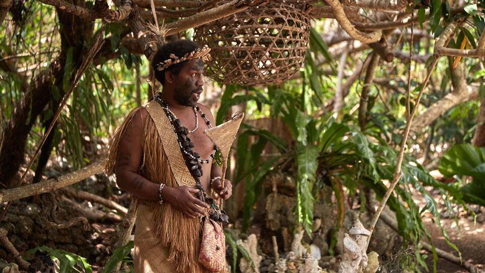 A person in traditional attire holding a woven basket and a small bag, standing in a forested area with dense foliage and tree branches overhead.