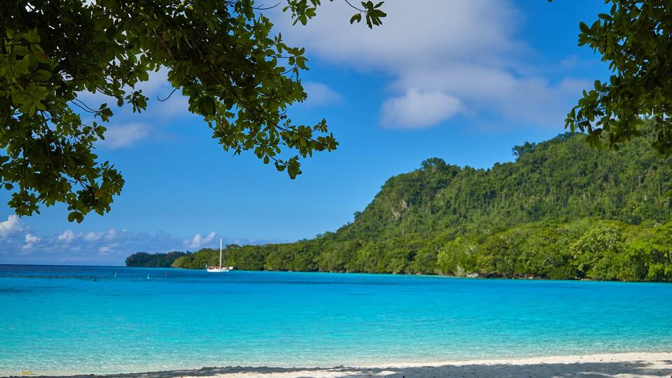 A beach with turquoise water, a sailboat, and a forested hill.