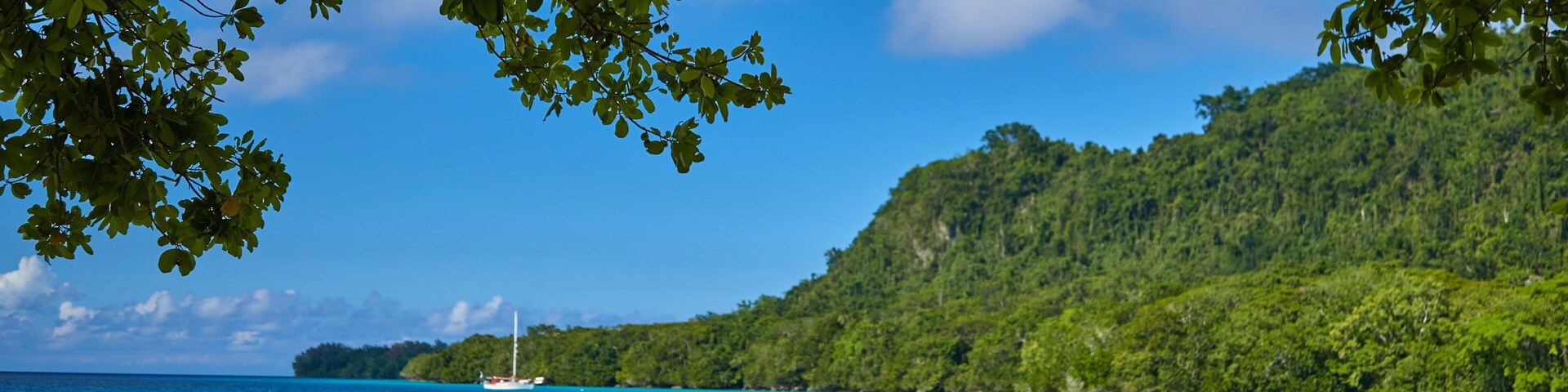 Uma praia com águas turquesa, um veleiro e uma colina arborizada.