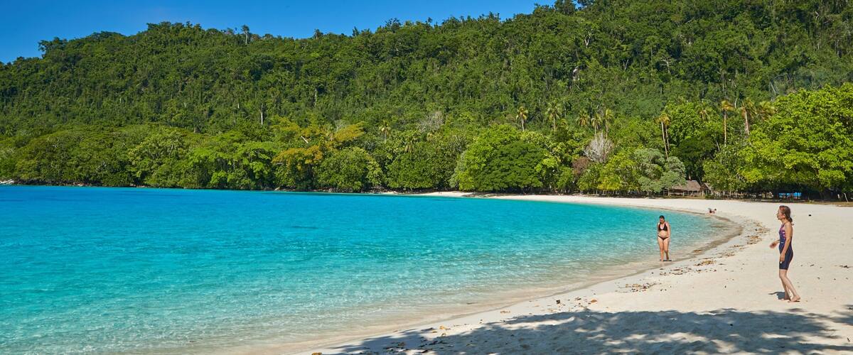 A beach with clear turquoise water, white sand, and a forested hill in the background.