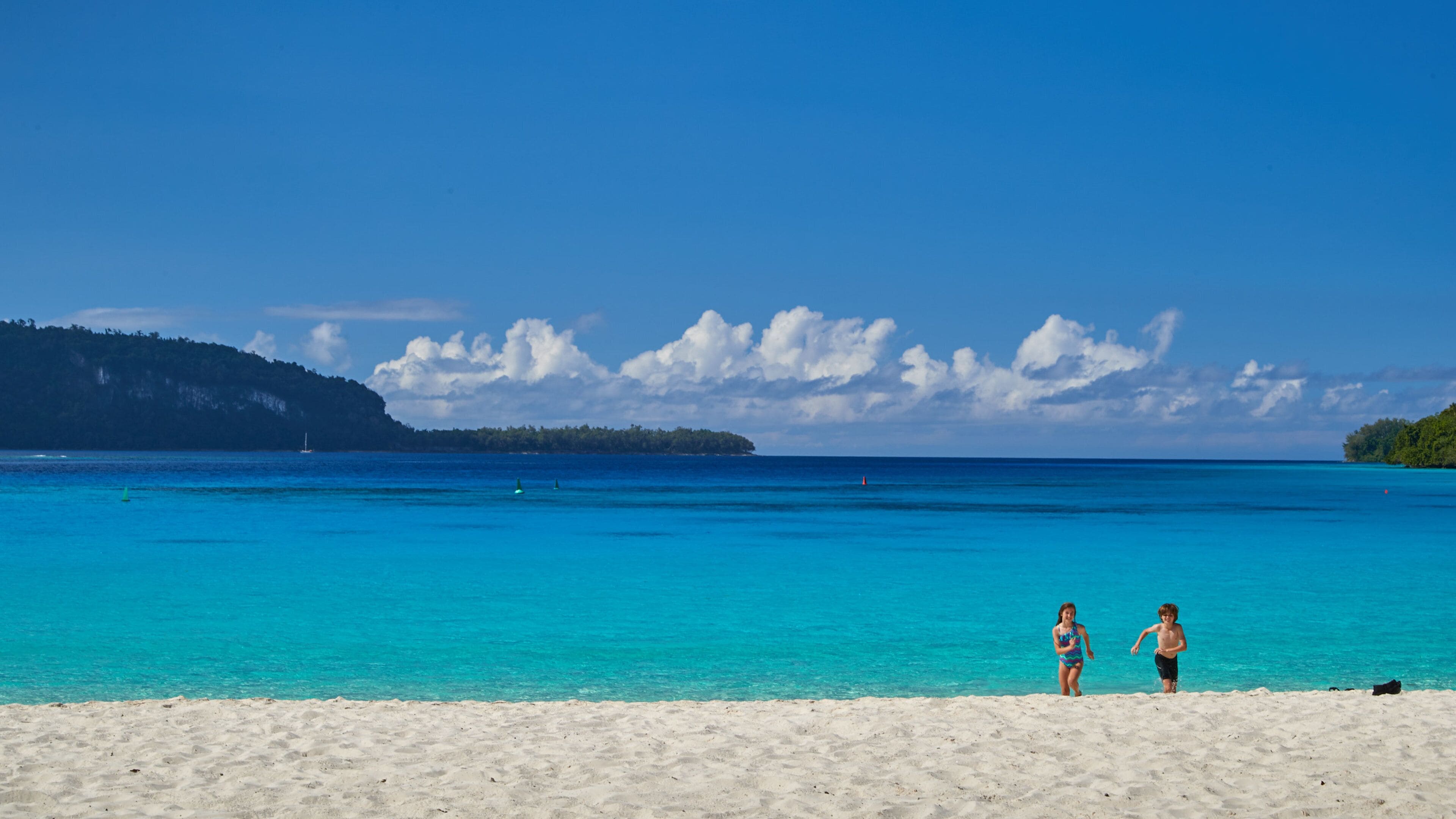 A beach with clear turquoise water, a sandy shore, and a distant island with a forested hill.