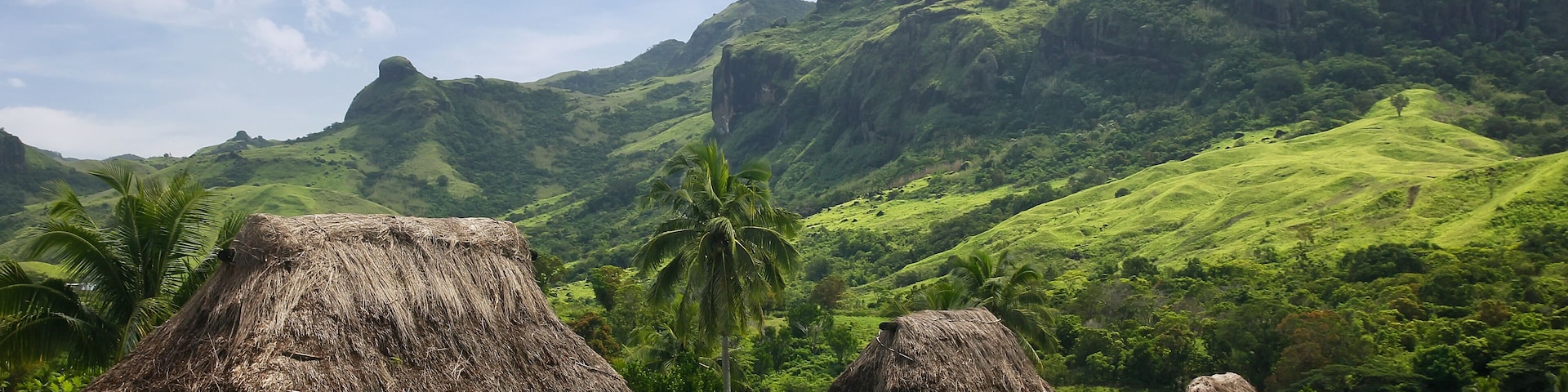 Cabanas com telhados de palha em paisagem verde exuberante, com colinas ao fundo.