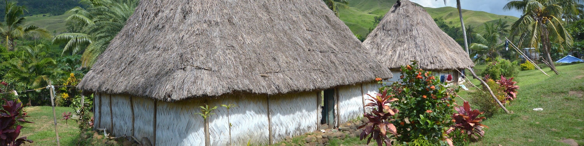 Dos chozas con techo de paja, un paisaje verde exuberante y palmeras al fondo.