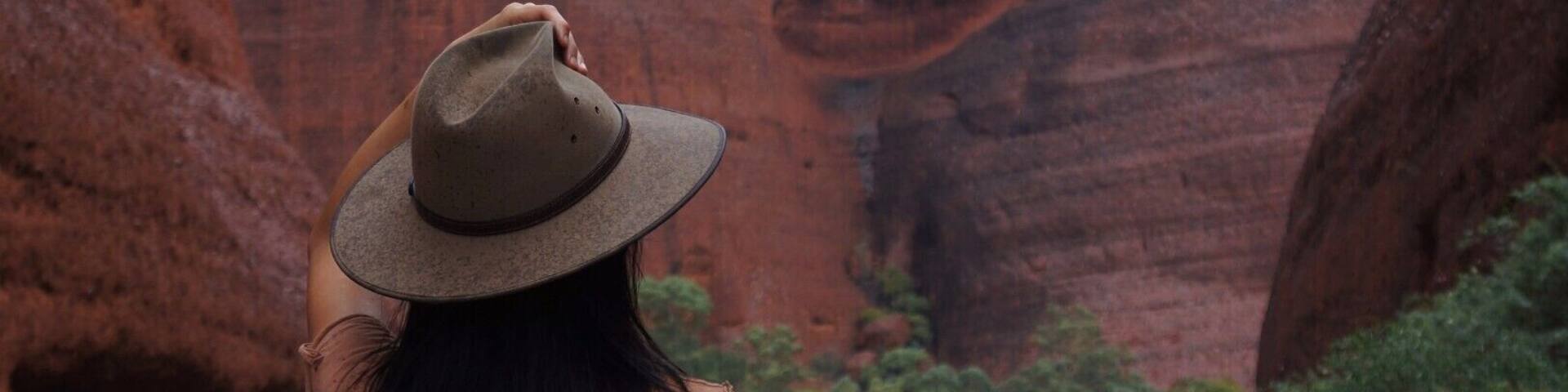 Una persona con sombrero y camisa marrón parada sobre un sendero rocoso, con formaciones rocosas rojas y vegetación al fondo.