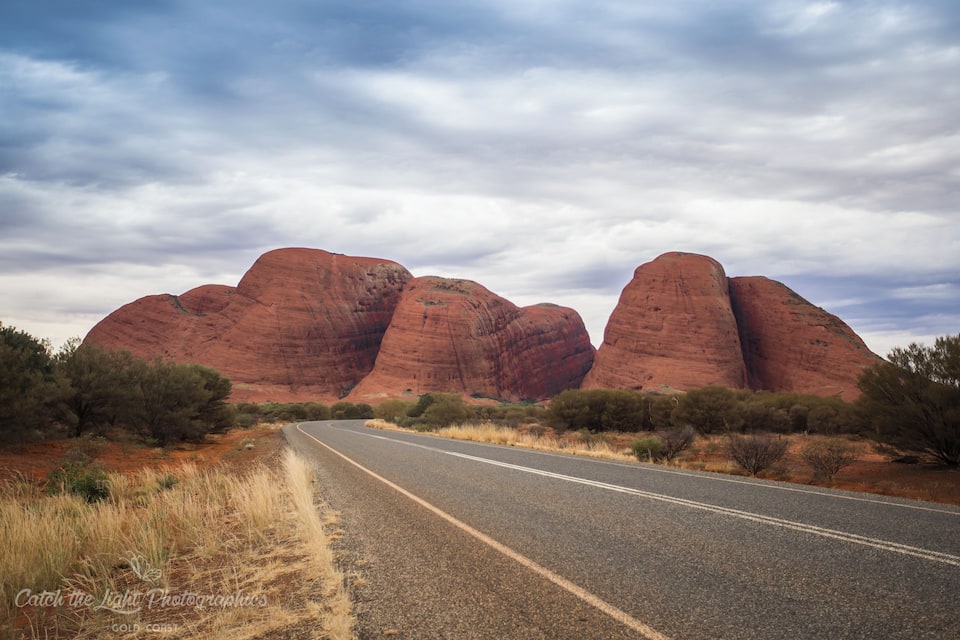 A road curving through a desert landscape with red rock formations in the background.