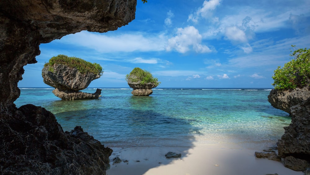 Una spiaggia con acque turchesi cristalline, due formazioni rocciose con vegetazione e cielo sereno.