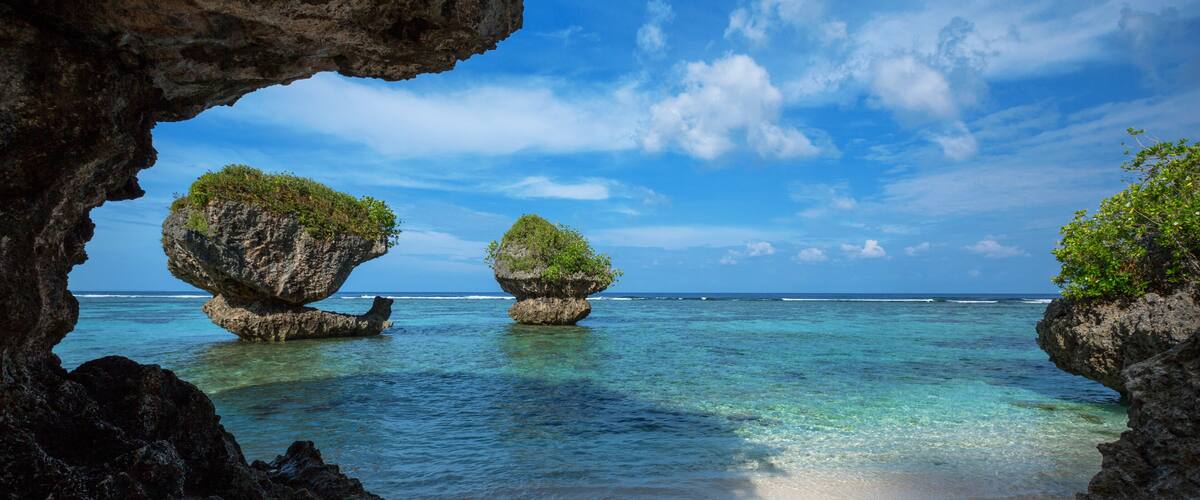 A beach with clear turquoise water, two rock formations with greenery, and a clear sky.