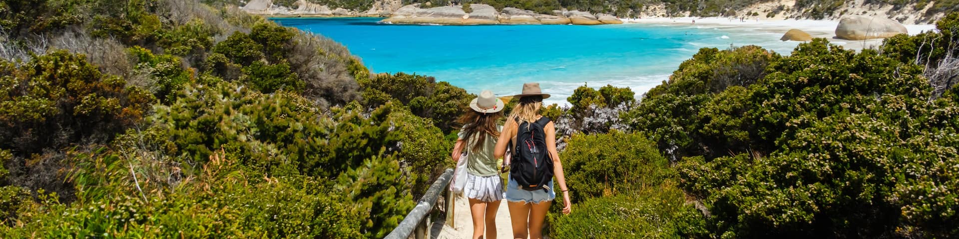 A coastal path leading to a beach with clear turquoise water, surrounded by lush greenery and rocky hills.