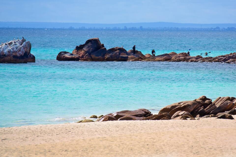 A beach with clear turquoise water, large rocks, and a distant city skyline.