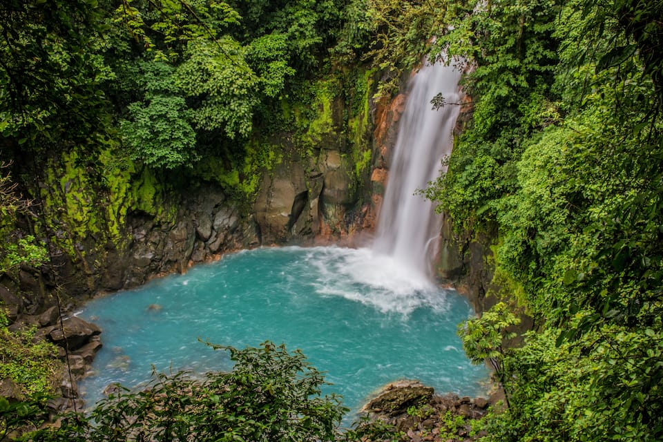 A turquoise pool surrounded by lush greenery and a waterfall.