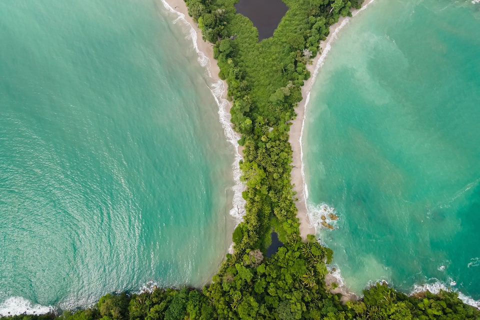 An aerial view of a beach with turquoise waters, dense green vegetation, and a small dark lake.