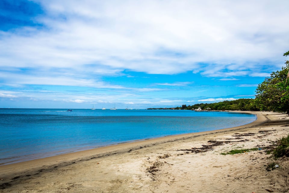 A sandy beach with clear blue water, boats in the distance, and trees along the shore.