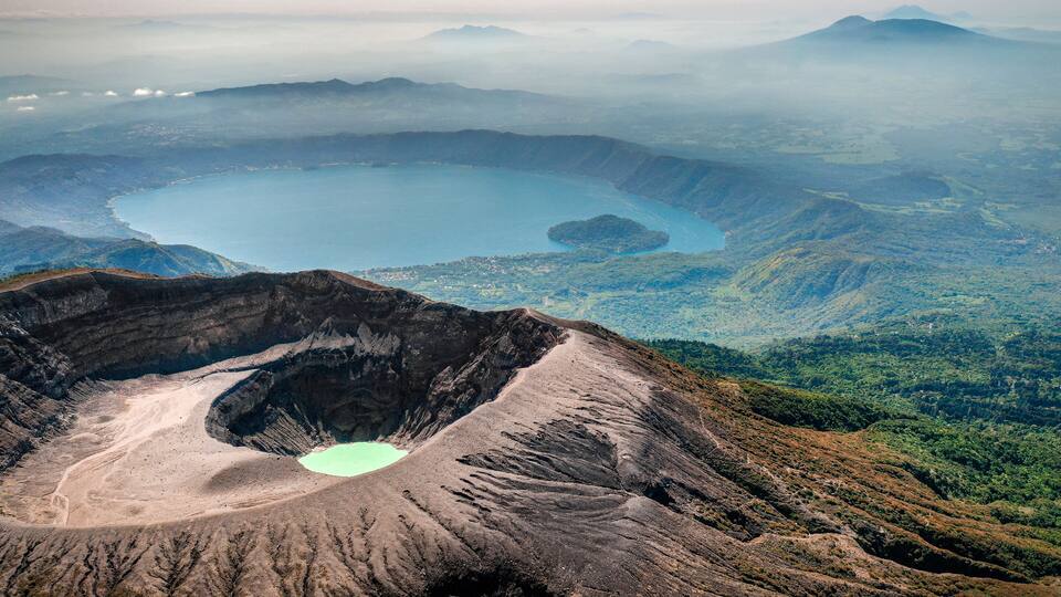 A volcanic crater with a lake, surrounded by mountains and forested areas.