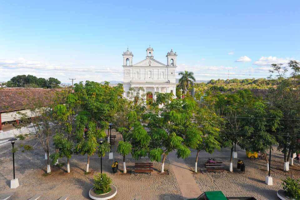 Una iglesia blanca con dos torres, rodeada de árboles y un espacio pavimentado con bancos.