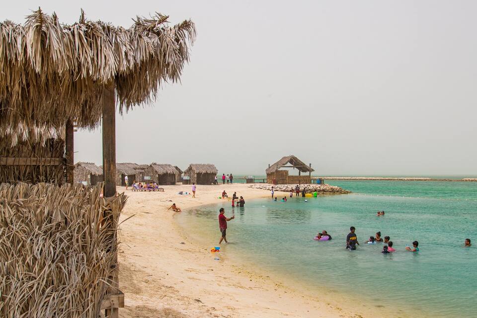 A beach with thatched huts, clear turquoise water, and people swimming and relaxing.
