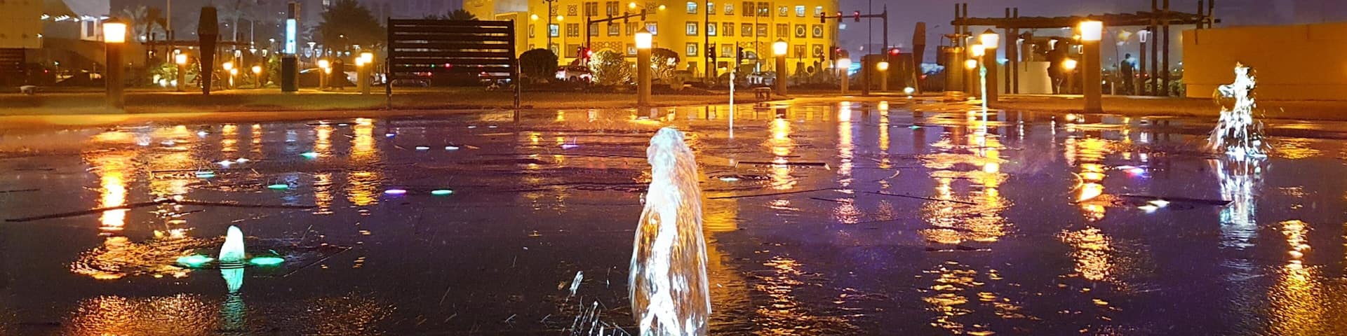 A night scene with a large illuminated building with a tower, a reflecting water body, and street lamps.
