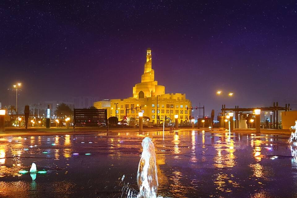 Escena nocturna amb un edifici gran il·luminat, una torre, un cos d'aigua que reflecteix i fanals.