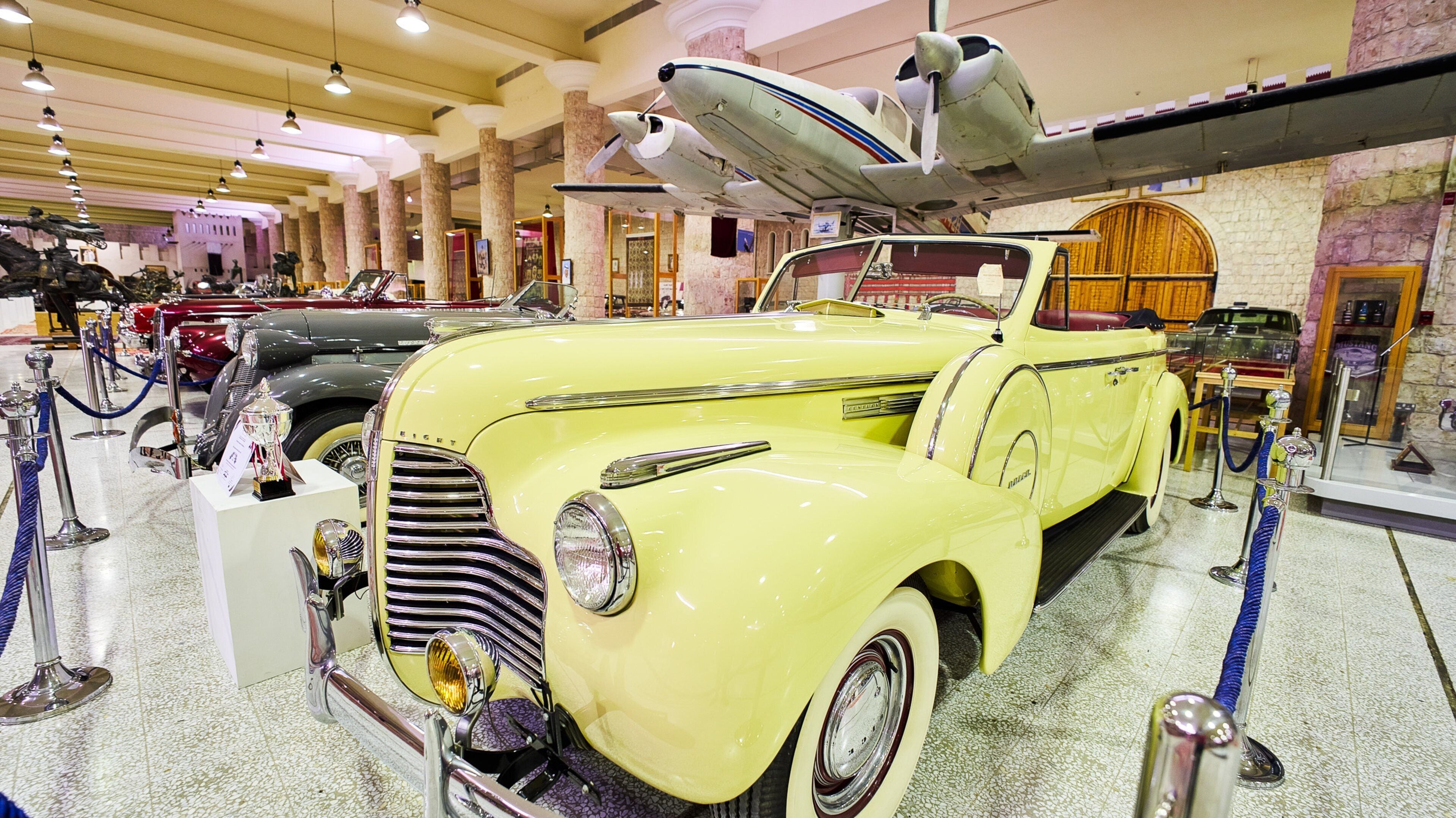 A vintage yellow car with a propeller attached to its roof, displayed in a museum with other classic cars and motorcycles.