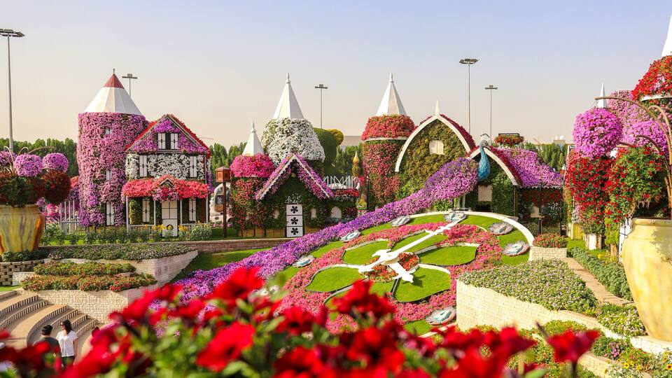 Um jardim com edifícios fantásticos cobertos de flores, um canteiro circular grande e um vaso amarelo em destaque.