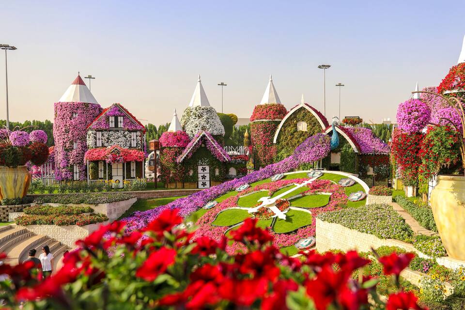 A garden with whimsical buildings covered in flowers, a large circular flower bed, and a prominent yellow planter.