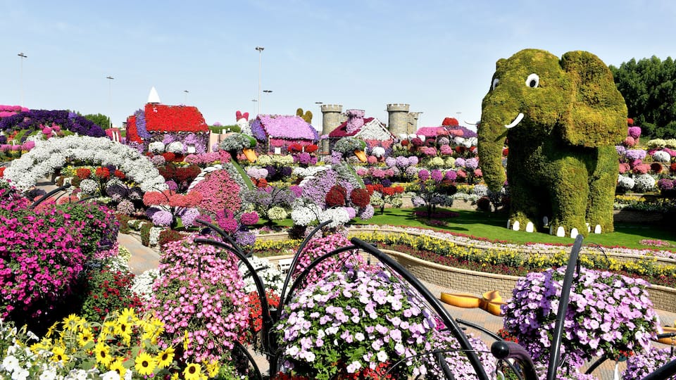 Um jardim com uma grande escultura de elefante, flores coloridas e uma estrutura que lembra um castelo.