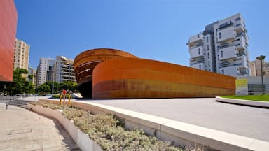 A modern curved building with a reddish-brown facade, surrounded by urban high-rise buildings and a clear blue sky.