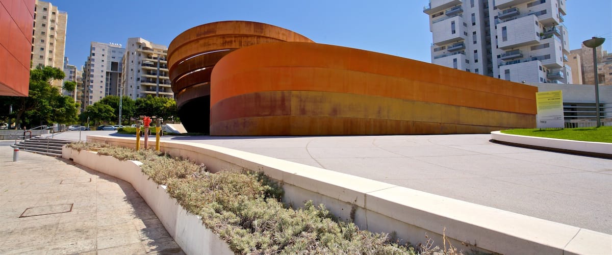 A modern curved building with a reddish-brown facade, surrounded by urban high-rise buildings and a clear blue sky.
