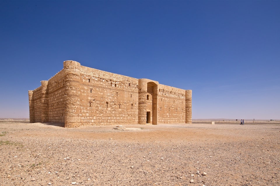 Un grand bâtiment ancien en pierre, doté d’un portail imposant, situé dans un paysage aride sous un ciel bleu dégagé.
