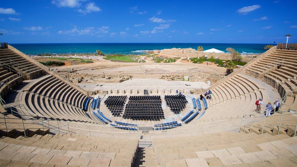 An ancient amphitheater with a stage and audience seating, overlooking a coastal area with ruins and the sea.