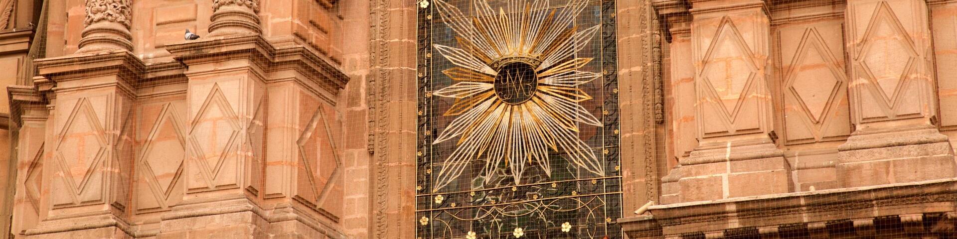 A detailed facade of a historic building with ornate carvings, statues, and a large stained glass window with a sunburst design.