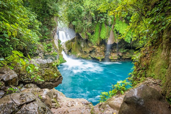 Ein Wasserfall stürzt in einen klaren, blauen Pool, umgeben von üppiger Vegetation und felsigem Gelände.