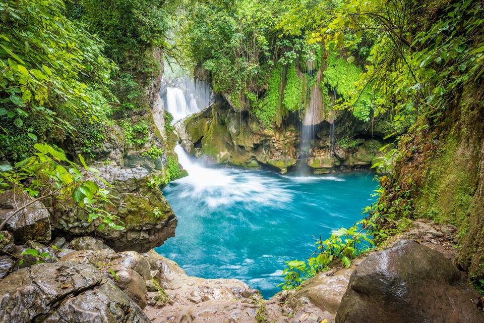 A waterfall cascading into a clear blue pool surrounded by lush greenery and rocky terrain.