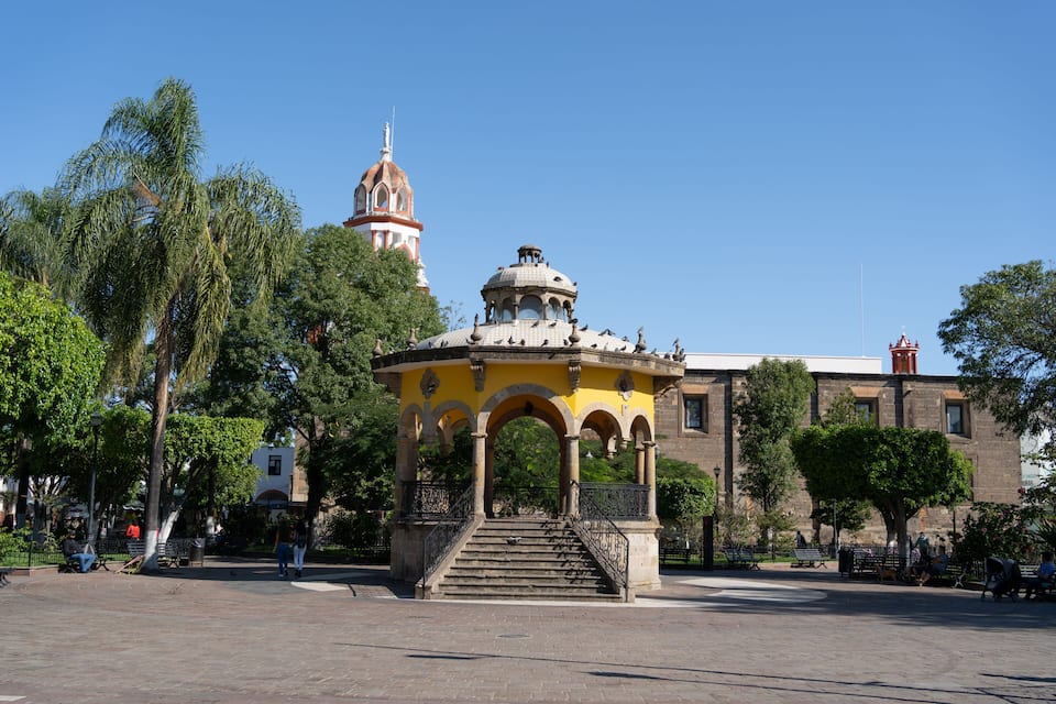 A historical plaza with a central fountain, a domed building in the background, and surrounding trees.