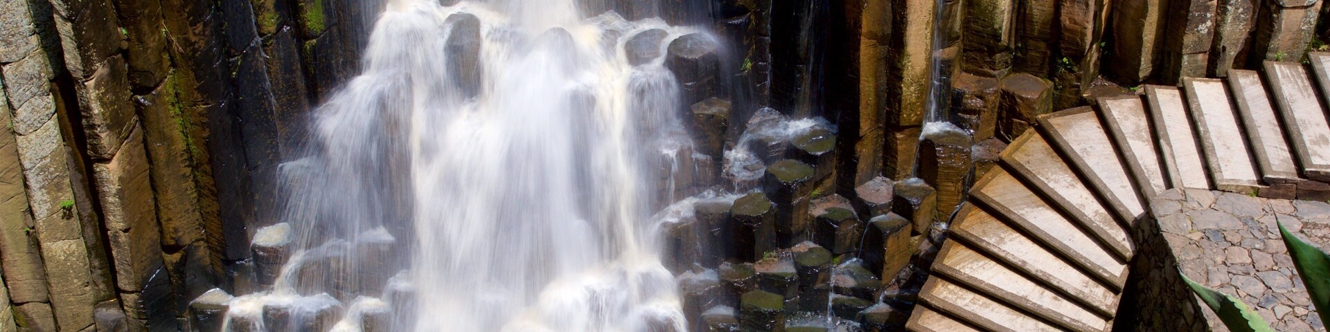 Una cascada fluye sobre rocas hacia un estanque, rodeado de escalones de piedra y vegetación.