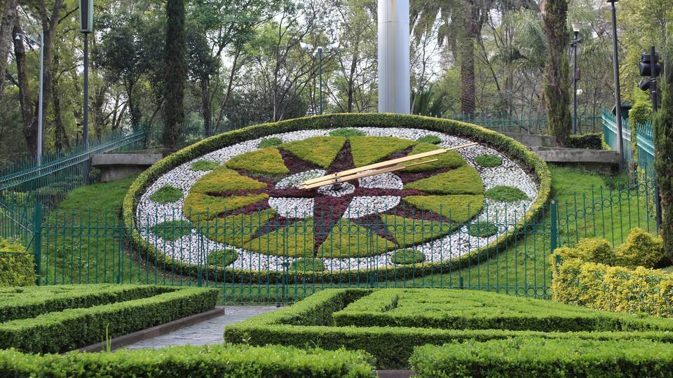 A meticulously landscaped garden with a central circular design featuring a star pattern, surrounded by neatly trimmed hedges and a green metal fence.