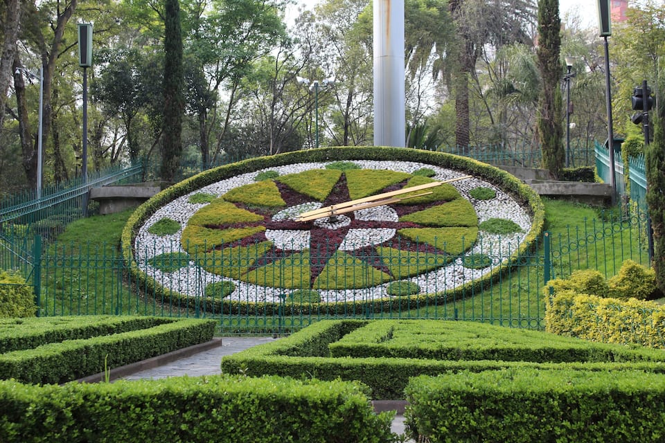 A meticulously landscaped garden with a central circular design featuring a star pattern, surrounded by neatly trimmed hedges and a green metal fence.