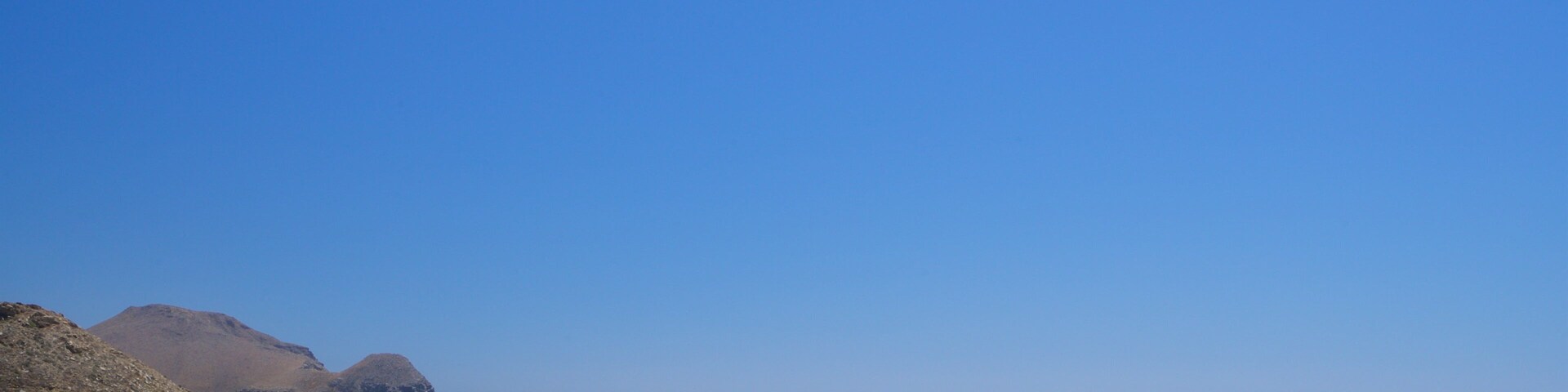 Uma praia com céu azul límpido, areal arenoso e colinas rochosas ao fundo.