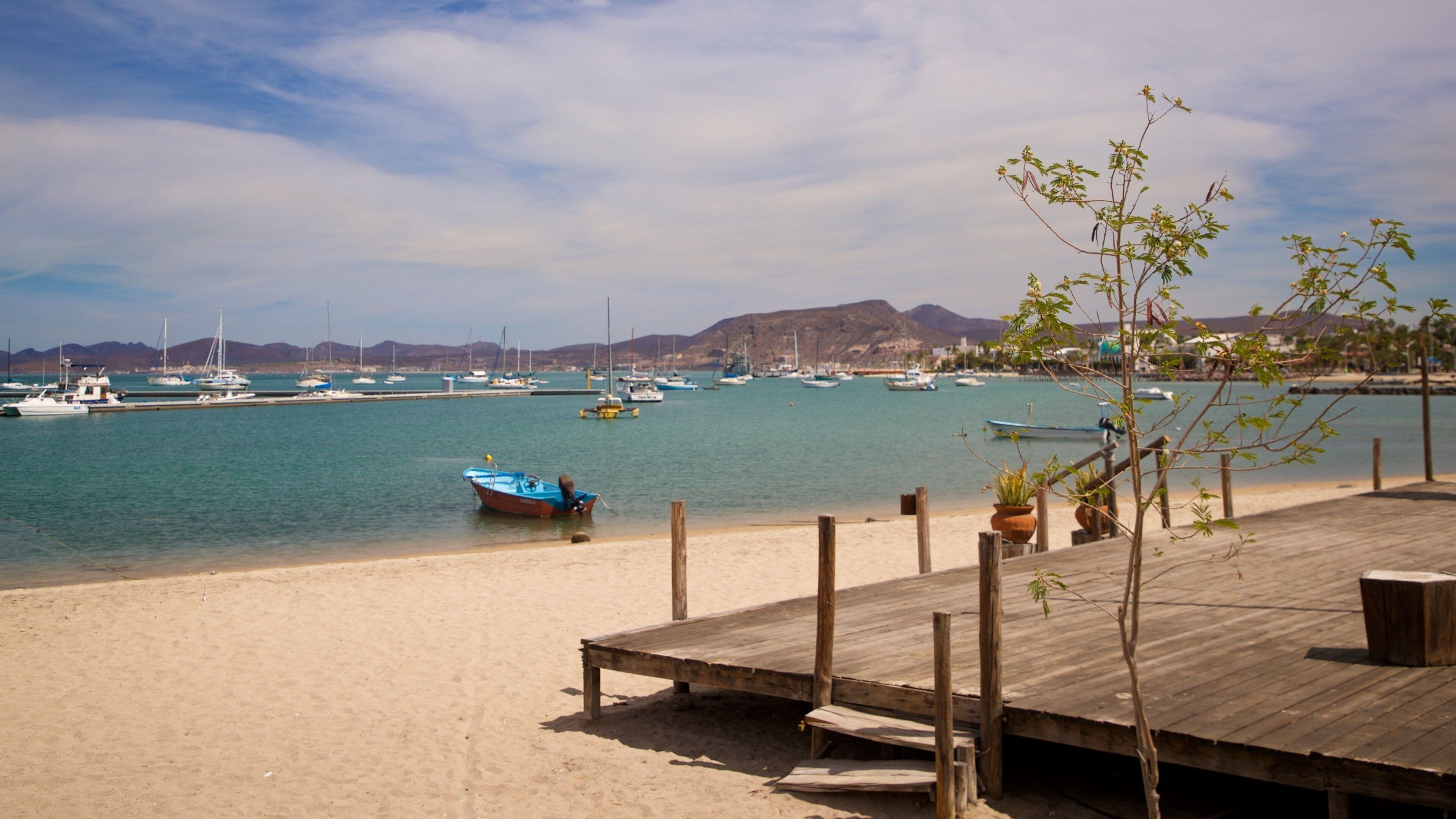 Una playa con un muelle de madera, barcos y montañas al fondo.