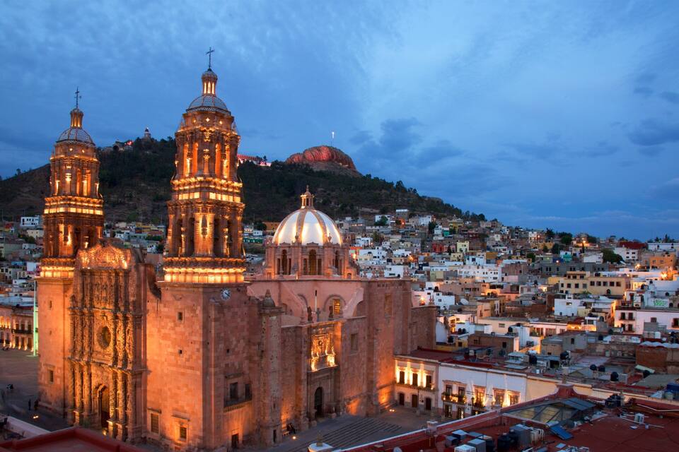 A historic cathedral with two towers and a dome, surrounded by a densely packed cityscape at dusk.