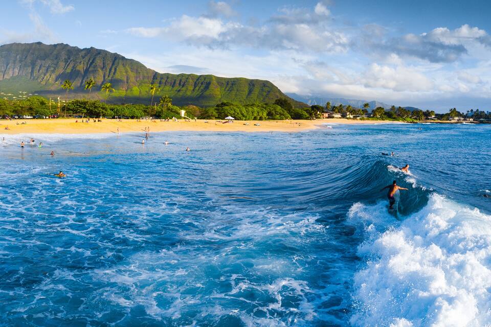 Une plage où l’on peut faire du surf, une montagne en arrière-plan et une eau d’un bleu limpide.
