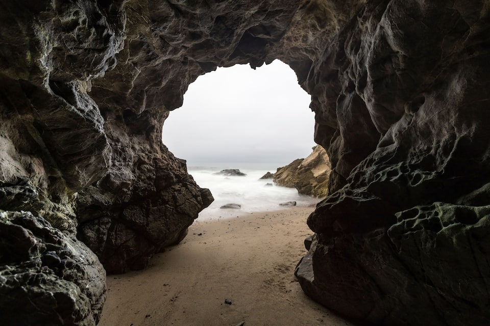 A rocky cave entrance with a view of the beach and ocean.