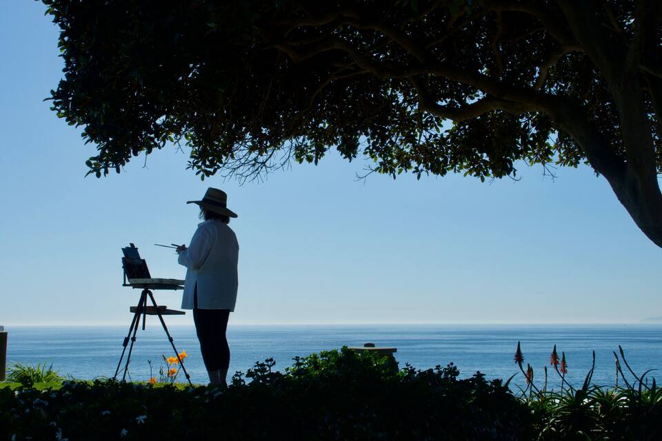 Una persona pintando junto al mar, bajo un árbol.